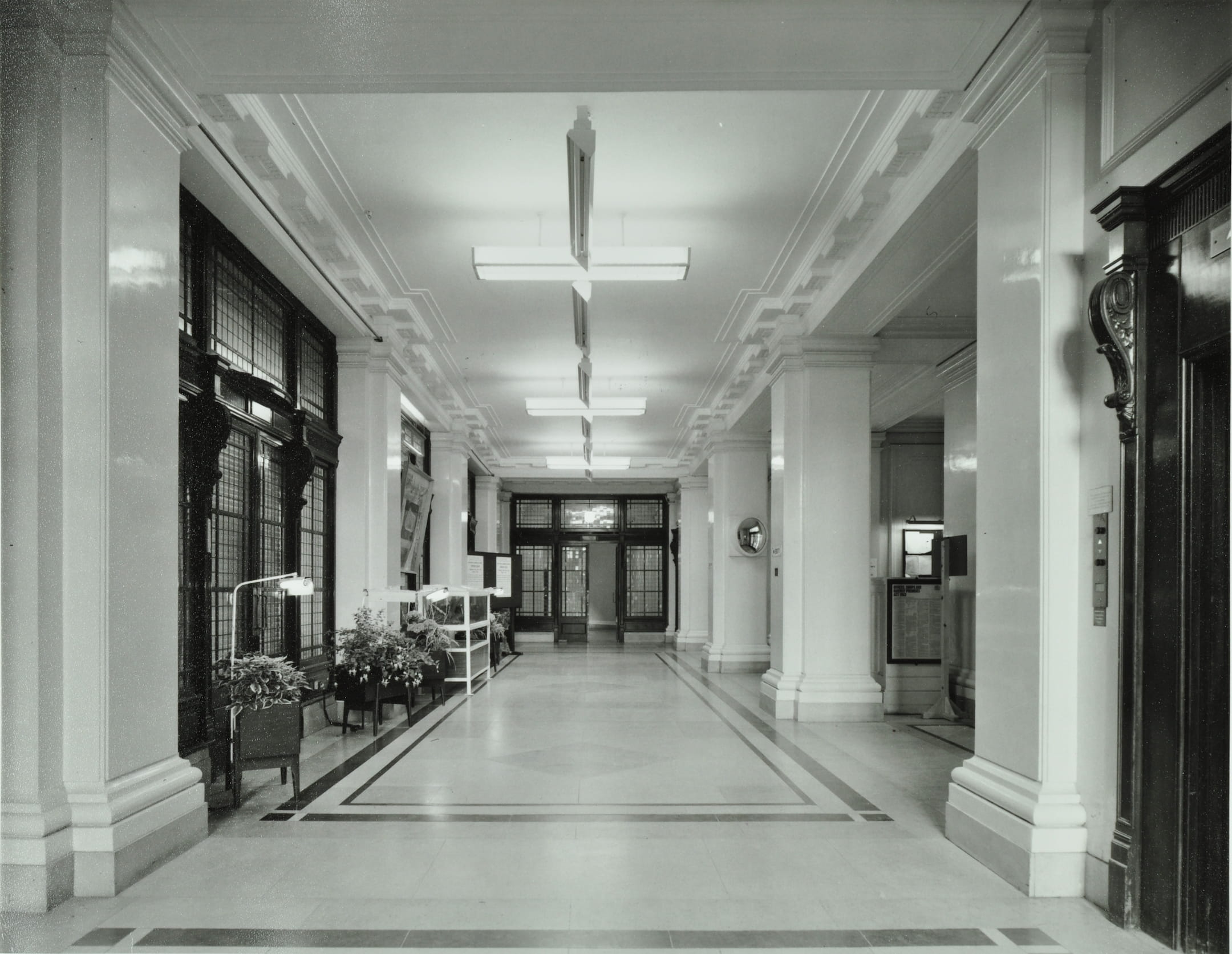 a long hall with stout columns and a staircase at the far end. The floor is stone. A noticeboard and some planters.