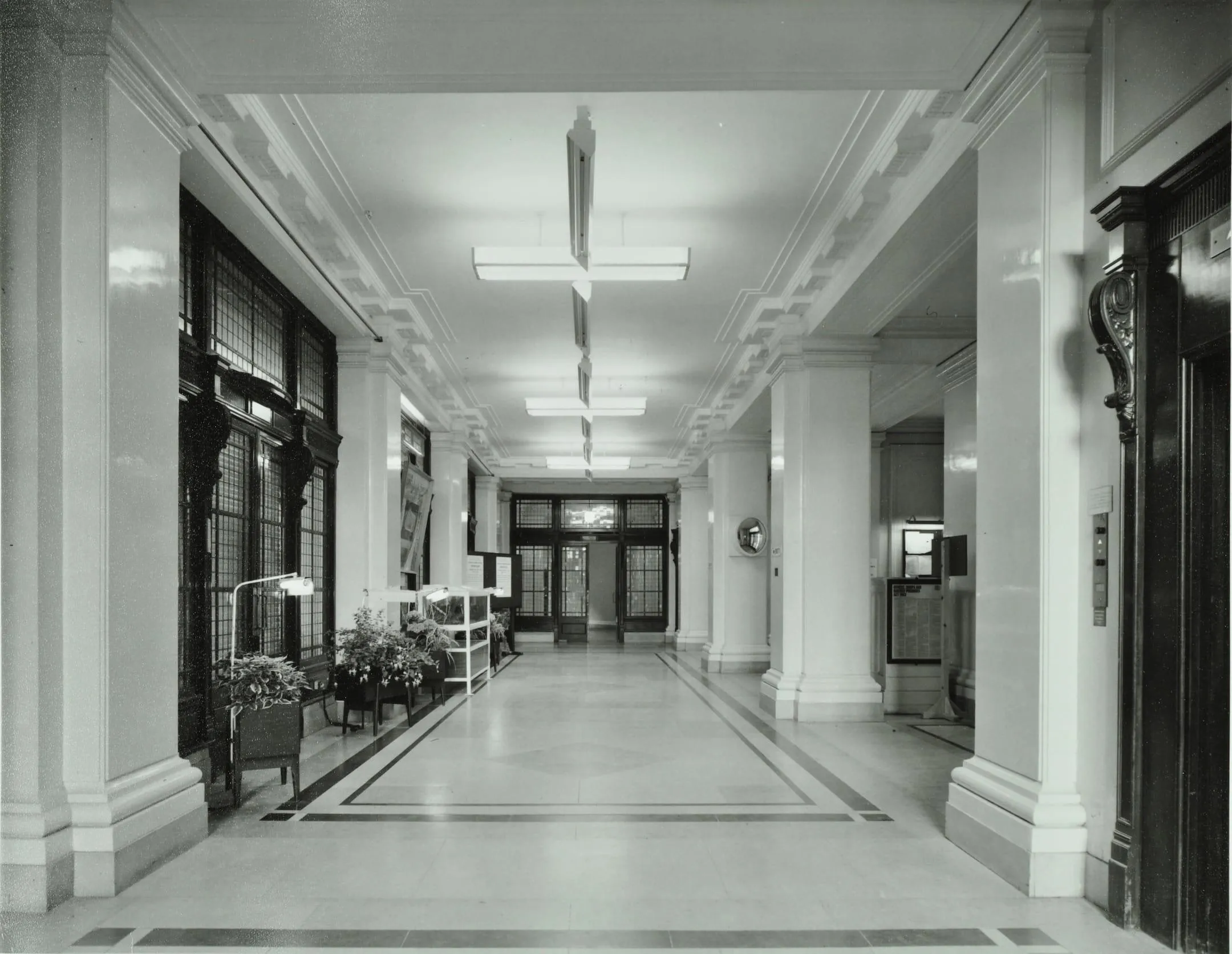 a long hall with stout columns and a staircase at the far end. The floor is stone. A noticeboard and some planters.