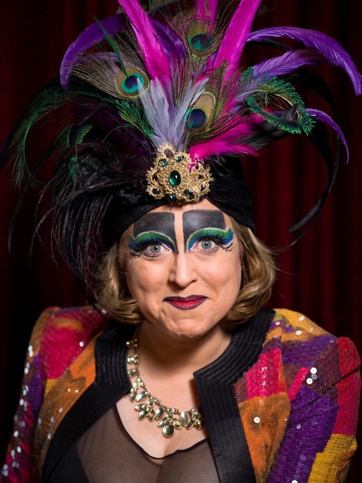 Portrait of a woman wearing bright orange, purple and yellow sparkley blazer, bold make up and feather hat 