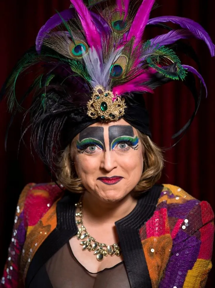 Portrait of a woman wearing bright orange, purple and yellow sparkley blazer, bold make up and feather hat