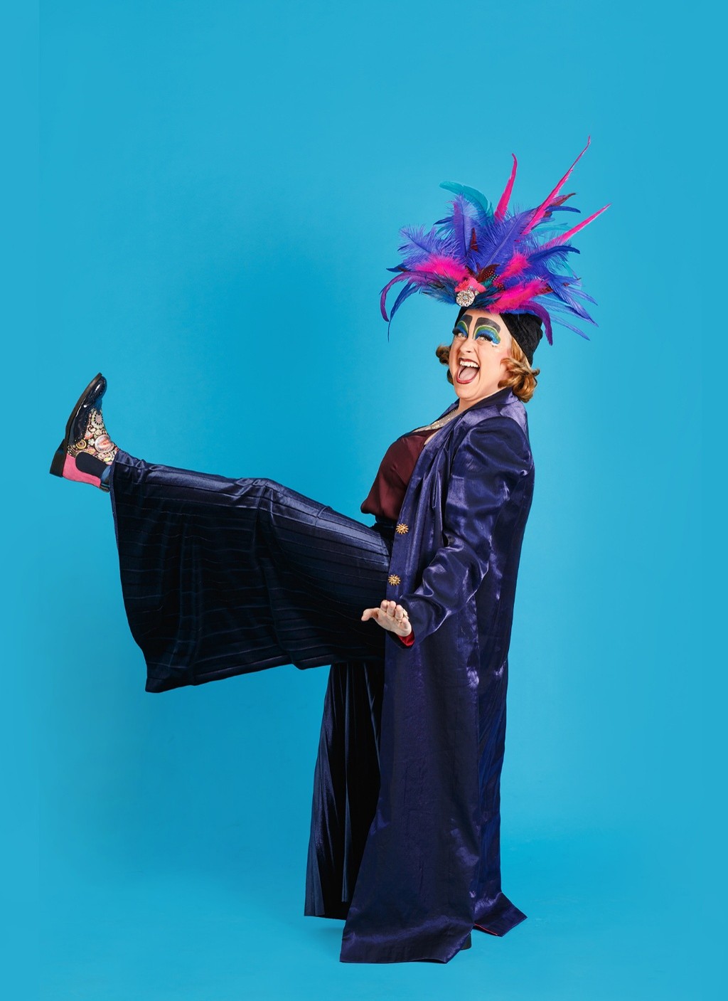 Woman in a purple clothes,  in front of a blue back drop with one leg front of her, a pink and purple feather hat. 