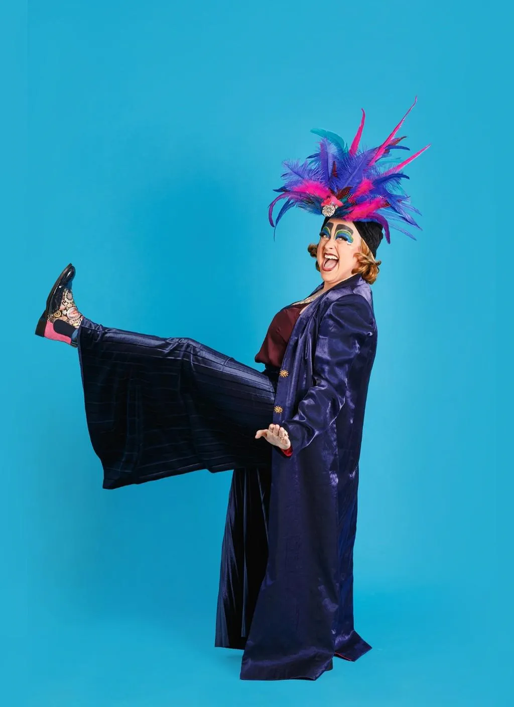 Woman in a purple clothes, in front of a blue back drop with one leg front of her, a pink and purple feather hat.