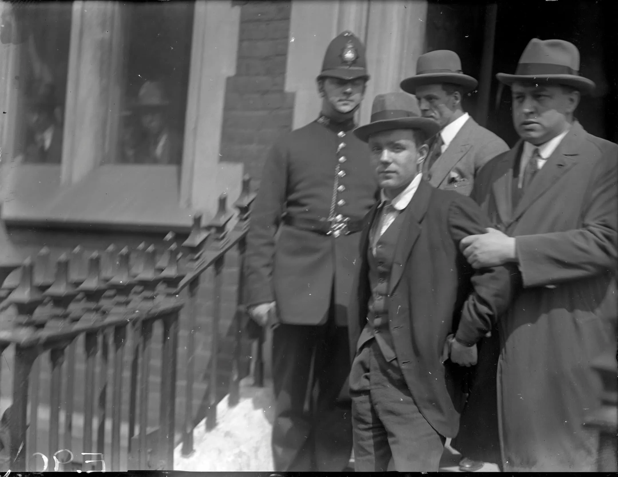 a man is held by the arms by a man in a trilby and a policeman watches over