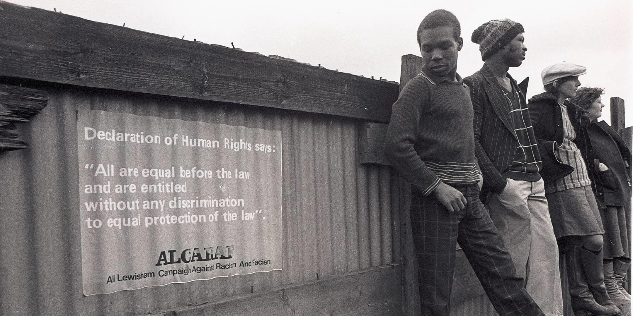 People leaning on a fence next to a poster with a declaration of human rights