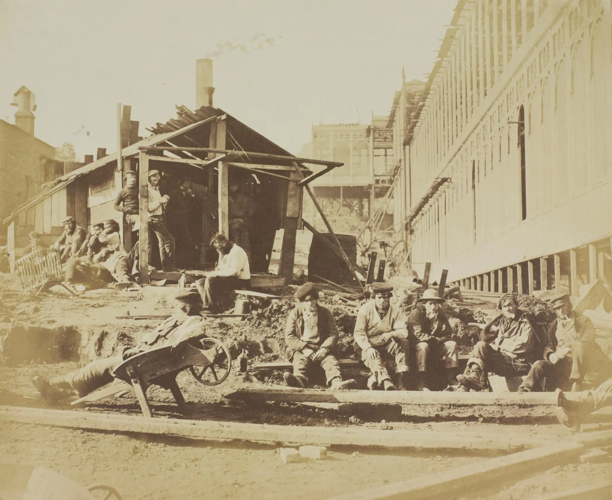 Photograph of workers taking a break for dinner, one man is in a wheelbarrow and the other sit near a wooden shack