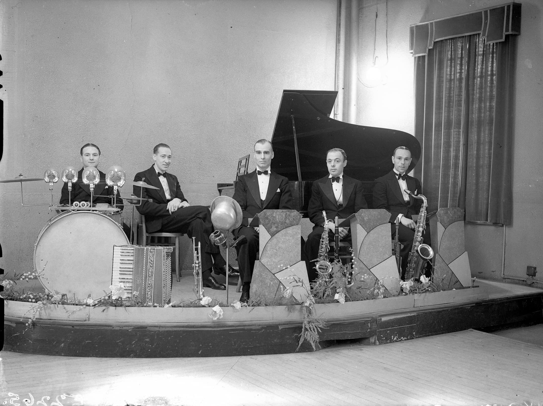 Group of five musicians, sitting on a stage, with their instruments, including a piano and drum
