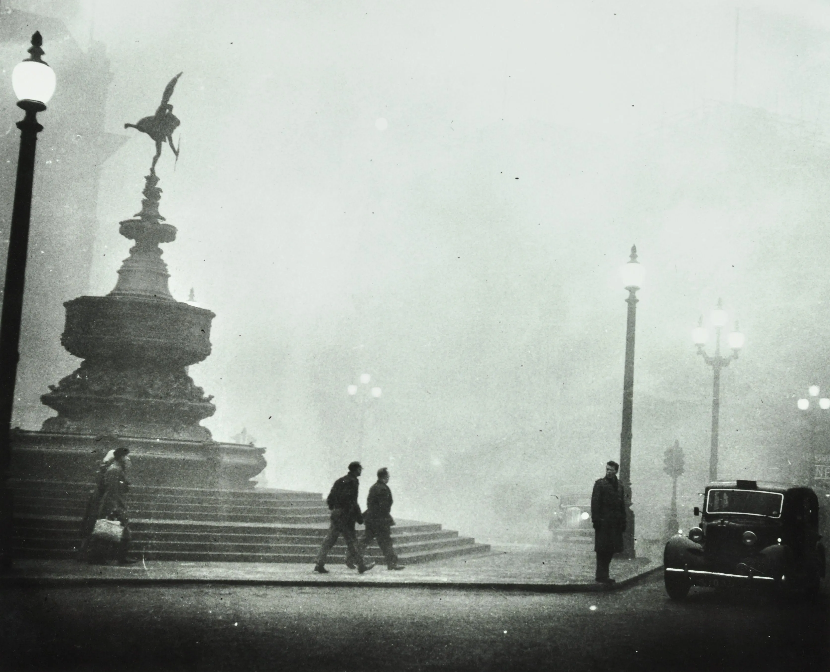 fog covers piccadilly circus as people walk by the eros statue and lamps are dimly lit