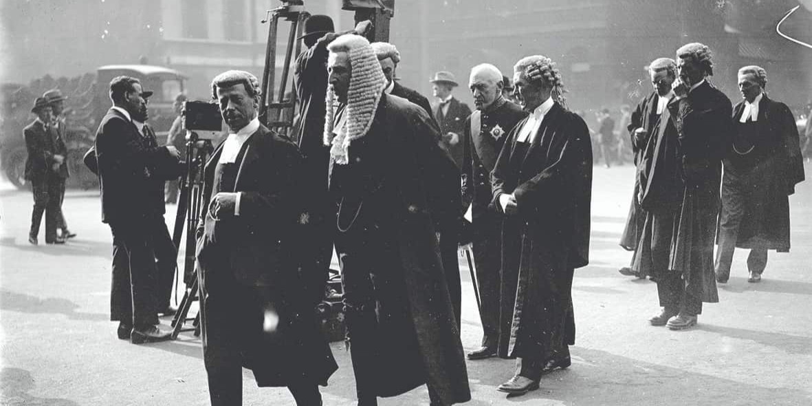 Judges and barristers walking in a group, news team behind them, crowds of people in the background