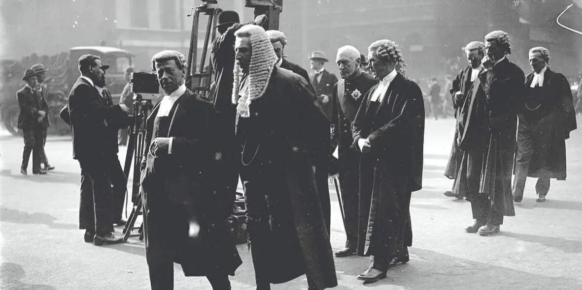 Judges and barristers walking in a group, news team behind them, crowds of people in the background