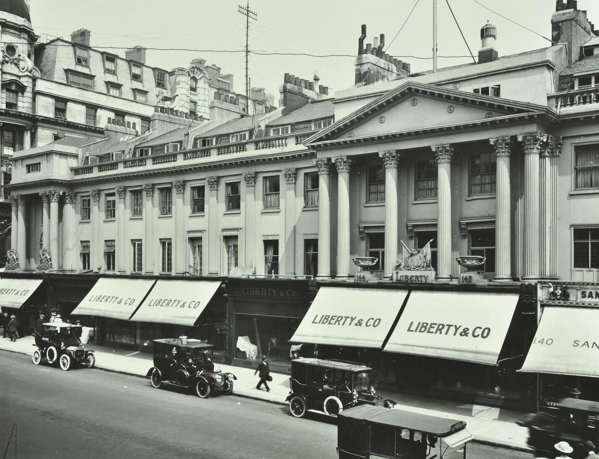 a classical building with shop front awnings with Liberty and Co. printed on. Old motor cars are parked up outside.