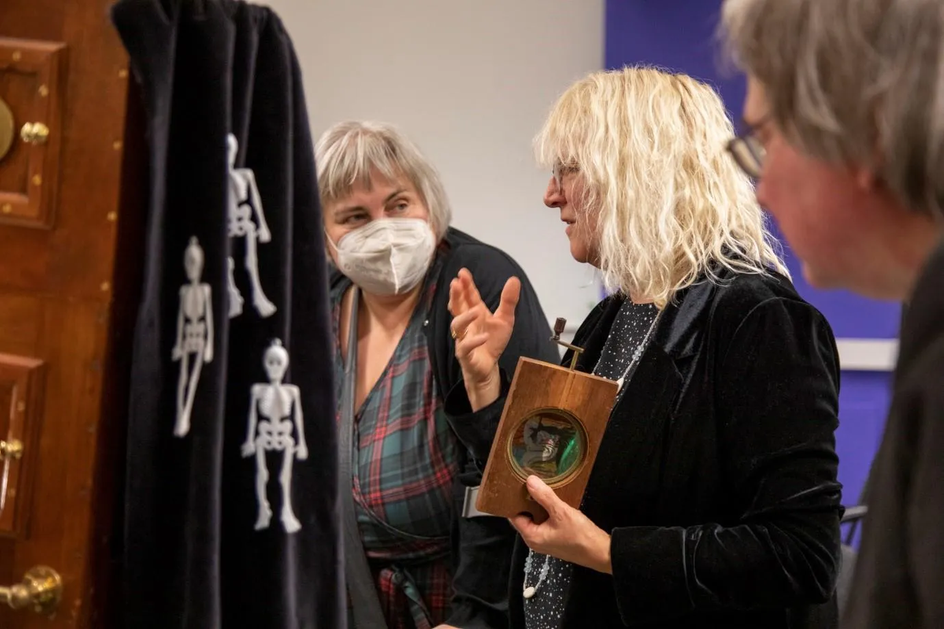 a woman holds a slide next to the magic lantern projector which has a skeleton cover draped over it