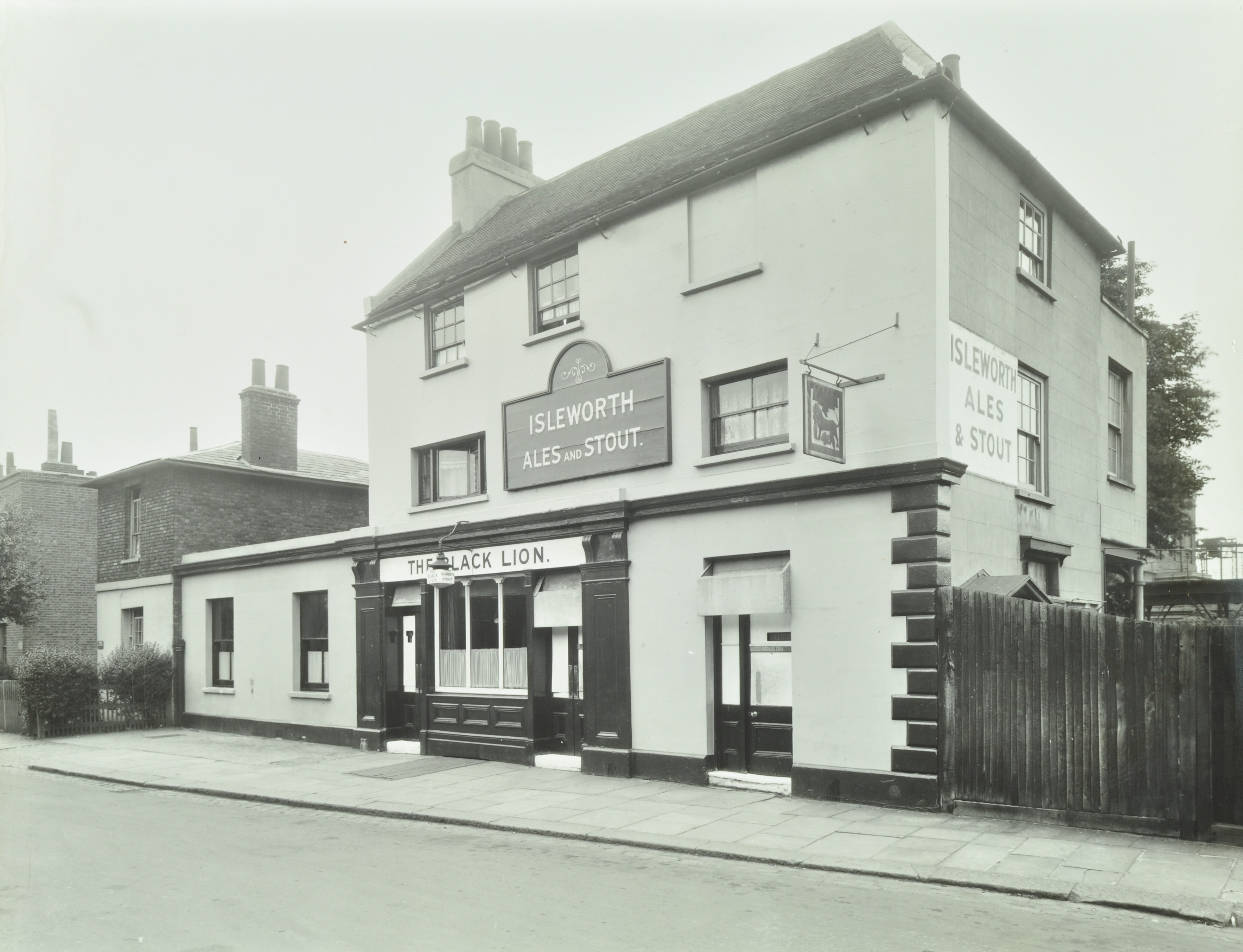 Outside of a pub, black doors, white sign that reads "The Black Lion", and another that reads "Isleworth Ales and Stout". 