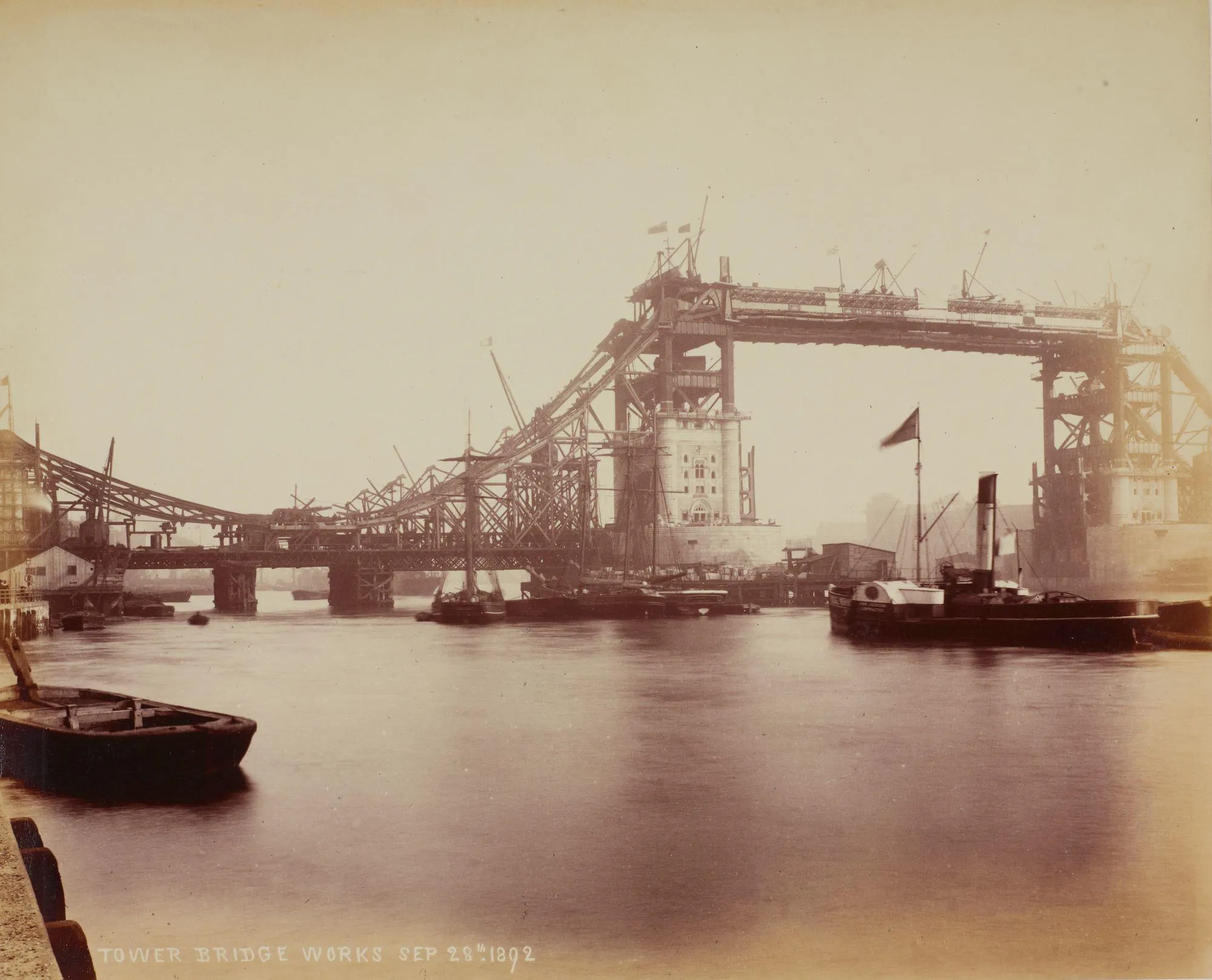 Tower Bridge being built, with scaffolding around the construction, and boats on the river